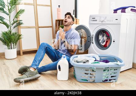 Junger hispanischer Mann, der schmutzige Wäsche in die Waschmaschine legt, bettelt und betet mit Händen zusammen mit Hoffnungsausdruck im Gesicht sehr emotional und Stockfoto