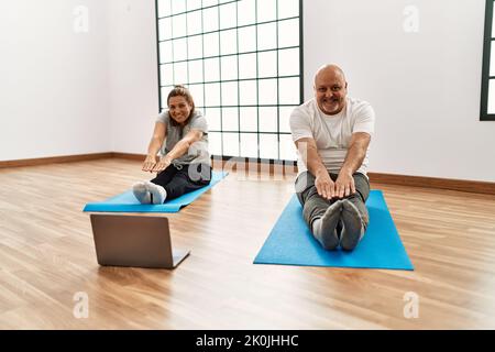 hispanische Paare im mittleren Alter, die sich mit einem Laptop im Fitnessstudio im Online-Sportunterricht ausstrecken Stockfoto
