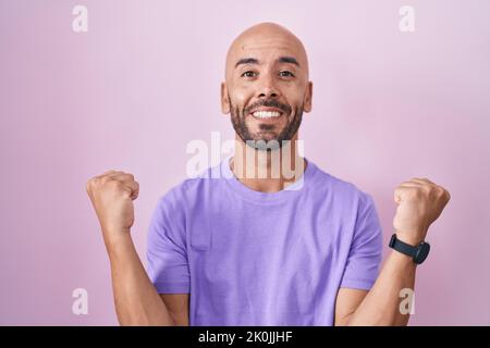 Kahlköpfiger Mann mittleren Alters, der auf rosafarbenem Hintergrund steht und stolz schreit, den Sieg und den Erfolg mit erhobenen Armen sehr aufgeregt feiert Stockfoto