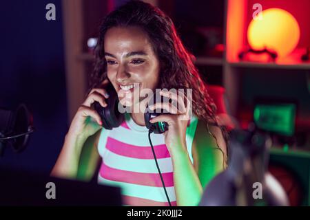 Junge schöne hispanische Frau streamer lächelnd zuversichtlich hält Kopfhörer im Spielsalon Stockfoto