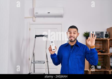 Hispanischer Reparaturman, der mit der Klimaanlage arbeitet, tut ein gutes Zeichen mit den Fingern, lächelt freundlich gestikuliert und ein ausgezeichnetes Symbol Stockfoto