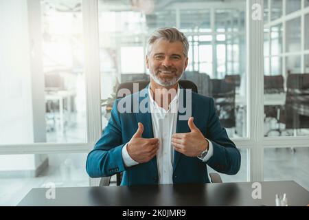 Lächelnder, reifer Geschäftsmann zeigt erhobene Finger auf, während er im Büro sitzt Stockfoto