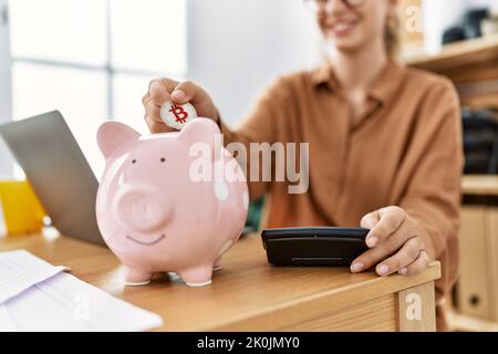 Junge blonde Frau lächelt selbstbewusst setzen Bitcoin auf Sparschwein im Büro Stockfoto