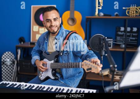 Junger hispanischer Musiker, der mit einem Lächeln auf den Lippen lächelt und im Musikstudio elektrische Gitarre spielt Stockfoto