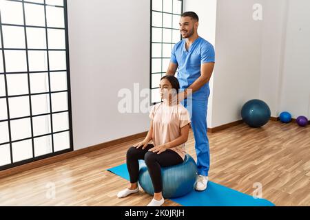Lateinischer Mann und Frau tragen Physiotherapeutenuniform mit fitem Ball massierenden Hals im Reha-Zentrum Stockfoto