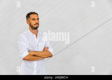 Hispanischer Mann in weißem Hemd und ernsthaftem Look mit gekreuzten Armen auf weißem Beton-Hintergrund. Stockfoto