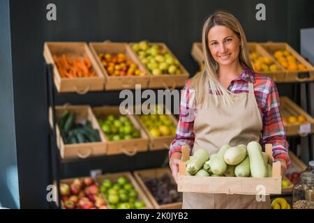 Frau arbeitet im Obst- und Gemüseladen. Sie hält den Korb mit Kürbis. Stockfoto