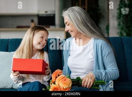 Fröhliche kaukasische kleine Mädchen genießt das Öffnen Geschenk-Box mit alten Dame mit Blumenstrauß im Wohnzimmer Stockfoto