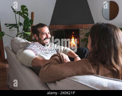 Lächelnder Mann und unkenntliche Frau sitzen zusammen auf dem Sofa und halten sich die Hände fest, während sie lachen und einander in der Wohnung mit Kamin anblicken Stockfoto