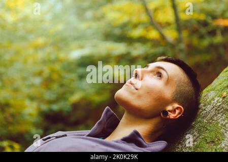 Junge Touristen, die in legerer Kleidung aufschauen, mit Rucksack, der auf dem Baum liegt und eine Pause einnimmt, um im herbstlichen Wald zu reisen Stockfoto