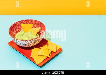 Knusprige Tortilla-Chips, die auf einem roten Teller in der Nähe der Schüssel mit einer leckeren Guacamole-Sauce auf blauem und gelbem Hintergrund platziert werden Stockfoto