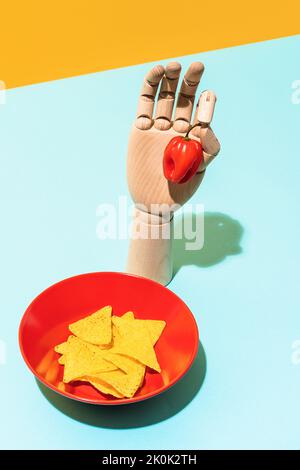 Von oben hölzerne künstliche Hand mit rotem Habanero-Pfeffer in der Nähe einer Schüssel mit knusprigen Tortilla-Chips der mexikanischen Küche auf blauem und gelbem Backgrou platziert Stockfoto