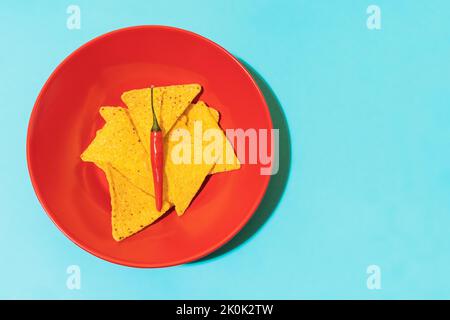 Draufsicht auf scharfe Chilischote auf knusprige Tortilla-Chips in roter Schüssel auf blauem Hintergrund Stockfoto