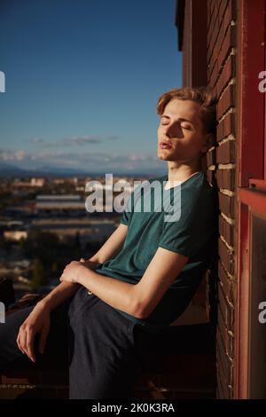Süßer junger blonder Mann, der sich an sonnigen Tagen mit geschlossenen Augen auf dem Dachrand ausruhte und sich gegen die Ziegelmauer lehnte Stockfoto