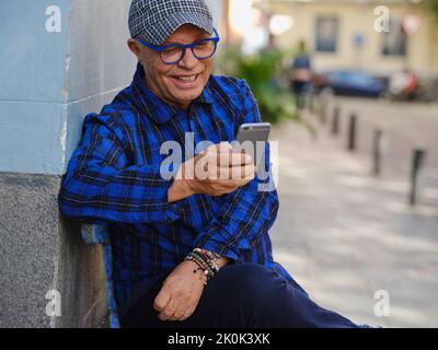 Hübscher Rüde im karierten Hemd und stylischen Brillen, die auf der Bank in der Stadt sitzen und auf dem Handy surfen Stockfoto
