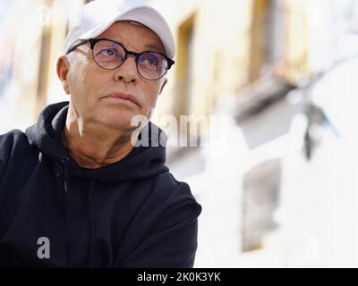 Gutaussehender älterer Mann in trendigen Brillen und stilvoller Kleidung, die auf verschwommenem Straßenhintergrund wegschaut Stockfoto