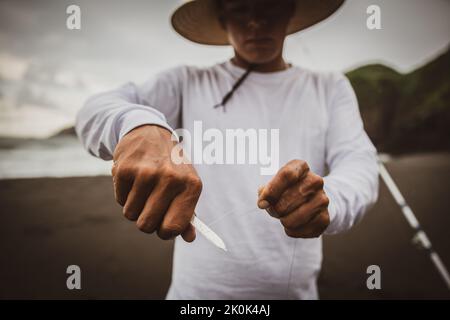 Crop anonyme Männchen in Hut schneiden Angelschnur mit Messer auf sandigen Ufer in der Nähe des Meeres, während die Vorbereitung für die Fischerei in der Natur Stockfoto