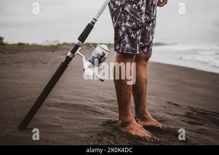 Barfuß-Männchen mit Angelrute in der Hand, der am Sandstrand in der Nähe des winkenden Meeres in der Natur steht Stockfoto