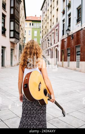 Rückansicht einer stilvollen lockigen Frau mit Gitarre, die durch ein helles Backsteingebäude in der Stadt läuft Stockfoto