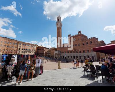 Piazza del Campo, ein berühmter mittelalterlicher Platz in der Stadt Siena, Toskana, Italien. Verkaufsstand mit Kleidung im Vordergrund und Restaurant rechts. Stockfoto