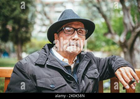 Ernst gealterter Mann mit schwarzem Kopfschmuck, der eine Brille trägt und wegschaut, während er auf einer Holzbank mit Zigarre in der Hand auf verschwommenem Hintergrund sitzt Stockfoto
