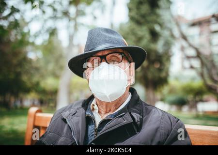 Gealterter Mann, der eine Schutzmaske mit Hut trägt und auf die Kamera schaut, während er auf einer Holzbank im Park auf unscharfem Hintergrund sitzt Stockfoto