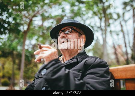 Von unten fröhlicher älterer Mann mit Schnurrbart, der eine Zigarre trägt und auf die Kamera schaut, während er auf der Bank im Park mit grünen Bäumen auf Blurr sitzt Stockfoto