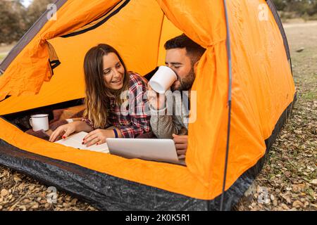 Junges Paar, das im farbenfrohen Zelt zeltet, Kaffee trinkt, Buch liest und auf dem Laptop stöbert Stockfoto
