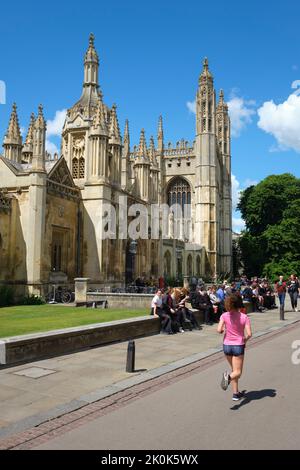 Sitzende Menschen und eine Joggerin, die an der unglaublichen Steinfassade des King's College vorbeikommt. In Cambridge, England, Vereinigtes Königreich. Stockfoto