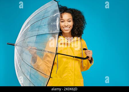 Afroamerikanische Frau in gelber wasserdichter Regenjacke mit transparentem Regenschirm isoliert auf blauem Studiohintergrund. Konzept der Herbstsaison Stockfoto