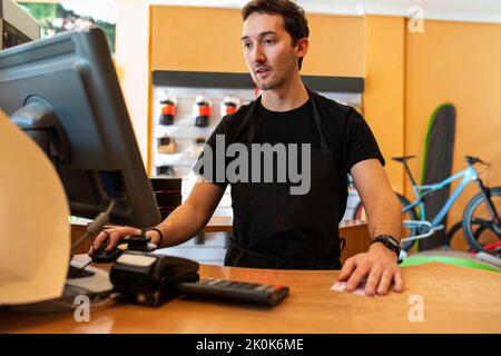 Fröhlicher junger Kassierer in schwarzem T-Shirt lächelnd und mit Computern während der Arbeit in einem modernen Fahrradladen Stockfoto