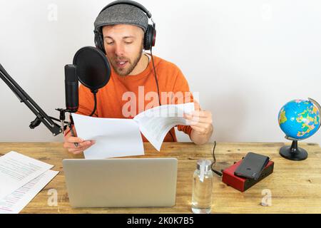 Konzentrierter Mann mit Kopfhörern, der Informationen aus dem Papier liest, während er im Sendestudio am Schreibtisch mit Laptop und Globus Reiseaudio aufzeichnet Stockfoto