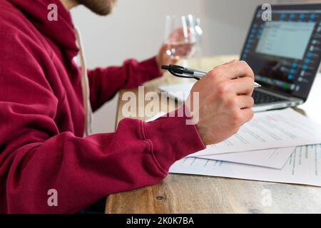 Nicht erkennbarer männlicher Arbeiter, der im Netbook im Internet surft und während der Arbeit im Lichtbüro mit einem Glas Wasser am Schreibtisch sitzt Stockfoto