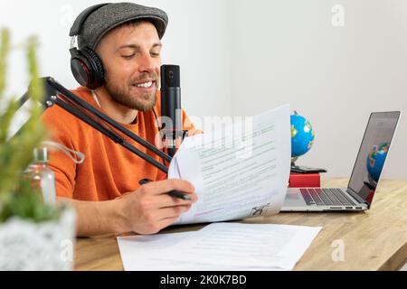 Konzentrierter Mann mit Kopfhörern, der Informationen aus dem Papier liest, während er im Sendestudio am Schreibtisch mit Laptop und Globus Reiseaudio aufzeichnet Stockfoto