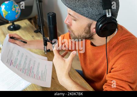 Konzentrierter Mann mit Kopfhörern, der Informationen aus dem Papier liest, während er im Sendestudio am Schreibtisch mit Laptop und Globus Reiseaudio aufzeichnet Stockfoto