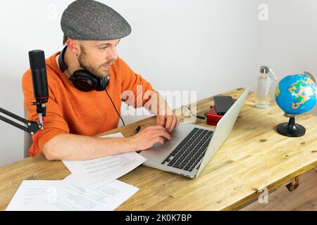 Konzentrierter Mann mit Kopfhörern, der Informationen aus dem Papier liest, während er im Sendestudio am Schreibtisch mit Laptop und Globus Reiseaudio aufzeichnet Stockfoto