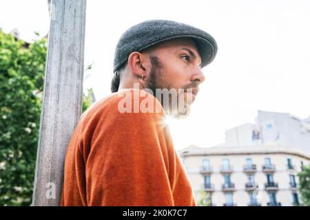 Von unten aus gesehen, wie ein bärtiger Mann mit trendiger Mütze neben dem Pfeiler auf der Straße steht und helles Sonnenlicht gegen Wohngebäude und Bäume hat Stockfoto