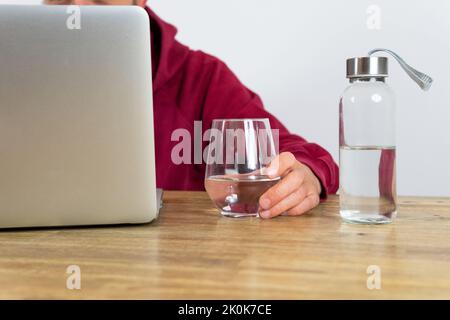 Ein nicht erkennbarer männlicher Arbeiter, der im Netbook im Internet surft, während er während der Arbeit im hellen Büro mit einer Flasche Wasser am Schreibtisch sitzt Stockfoto