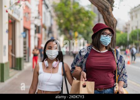 Junge peruanische Freundinnen in Freizeitkleidung und Schutzmasken, die während der Coronavirus-Pandemie in der Stadt auf der Straße in der Nähe von Gebäuden spazieren Stockfoto
