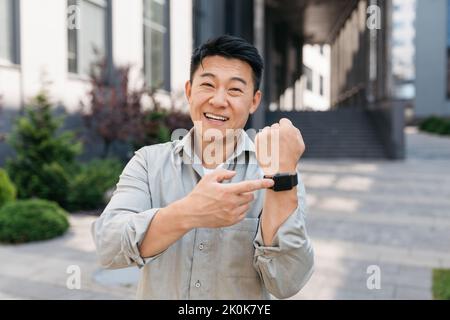 Porträt eines glücklichen asiatischen Geschäftsmannes, der mit dem Finger auf eine moderne Armbanduhr zeigt und vor die Kamera zeigt und im Freien steht Stockfoto