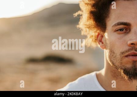 Halbgesichtsporträt eines ethnischen Mannes mit afrohaarigen Stehen auf einer unbefestigten Straße und Blick auf die Kamera am sonnigen Abend vor einem unscharfen felsigen Hügel Stockfoto