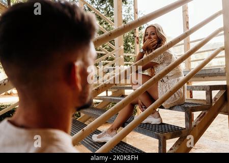 Von einer heiteren blonden Frau, die lächelt, einem anonymen Mann, der sich ansieht, während er auf einer Metalltreppe eines modernen Gebäudes gegen blu sitzt Stockfoto