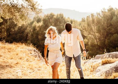 Fröhlicher Mann in modischer Freizeitkleidung und Frau in elegantem Kleid, die die Hände hält und wegschaut, während sie bei Sonnenuntergang auf einem Hügel zwischen Bäumen im Park spazieren geht Stockfoto