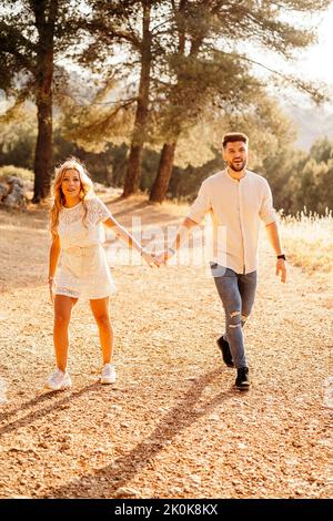 Fröhlicher Mann in modischer Freizeitkleidung und Frau in elegantem Kleid mit Hut, der die Hände hält und die Kamera anschaut, während sie auf einem Hügel zwischen Bäumen im Park spazieren geht Stockfoto
