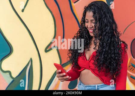 Content junge ethnische weibliche mit lockigen langen Haaren Textnachrichten auf dem Handy gegen Graffiti Wand Stockfoto