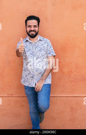 Entzückter ethnischer Mann, der wie eine Geste in der Nähe der Pfirsichwand in der Stadt stand und die Kamera ansah Stockfoto