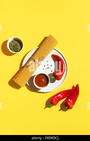 Von oben appetitlich frische trockene Spaghetti mit roter Sauce und Gewürzdip rote Paprika auf gelbem Hintergrund Stockfoto
