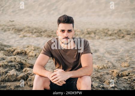 Seriöse gutaussehende bärtige hispanische Mann in braunem T-Shirt am Sandstrand, die Hände zusammenklappend und die Kamera anblickend Stockfoto
