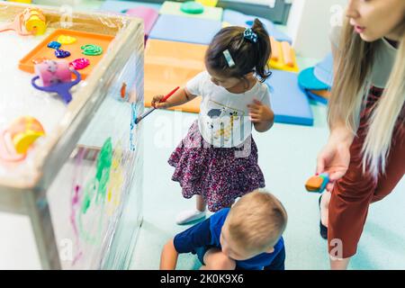 Frischhaltefolie für die Verbesserung der Phantasie von Kindern. Kleinkinder malen mit, Bürsten, Rollen und Farben auf einer Frischhaltefolie, die um den hölzernen Regalständer gewickelt ist. Lustige Aktivität für Kinder Sinnesfertigkeiten, Kreativität, räumliche Aufmerksamkeit und visuelle Wahrnehmungsentwicklung im Kindergarten. Hochwertige Fotos Stockfoto