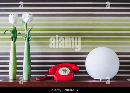 Blumen in grünen Vasen und Retro-Telefon neben Lampe in Reihe auf Holztisch gegen Retro-Tapete platziert Stockfoto